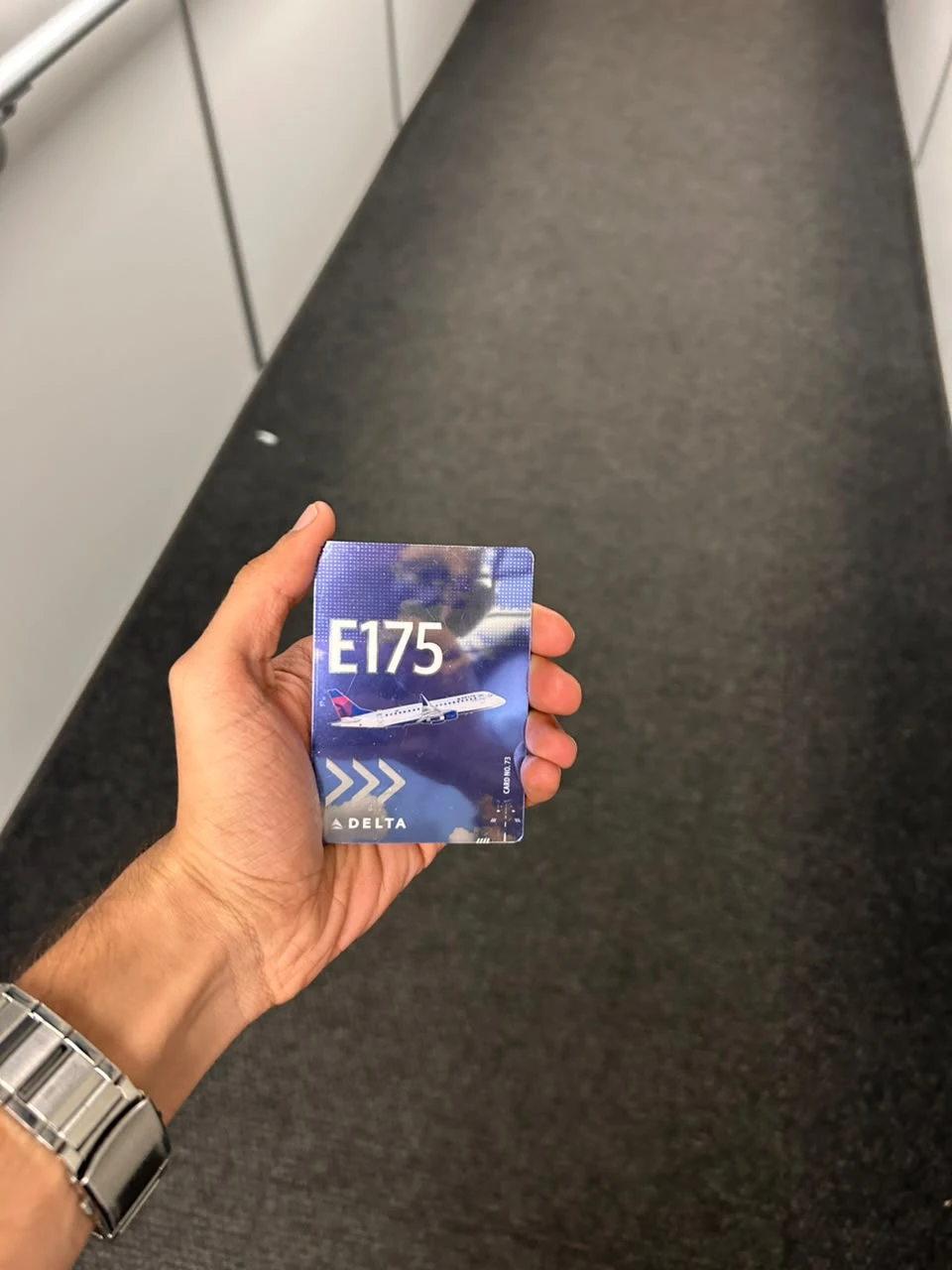 A holographic Delta trading card for the Embraer E175, card number 73, held in a hand in the aircraft aisle