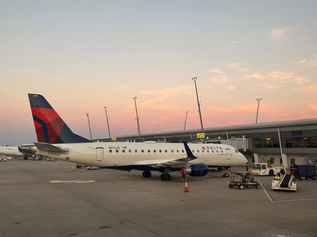 A Delta Connection Embraer E175 parked at gate A13 at Indianapolis International Airport under a pink sunset sky