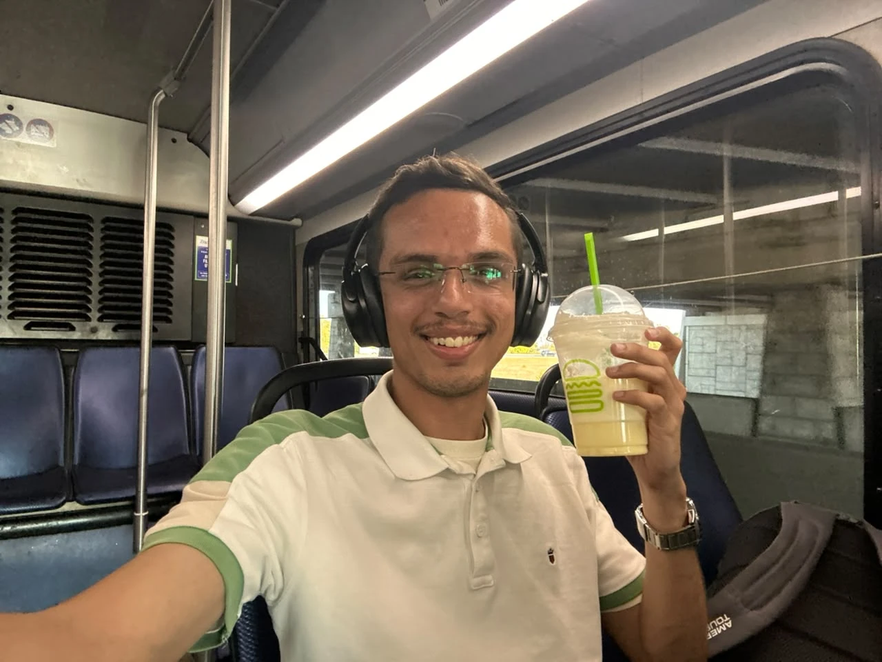 Aryan smiling on a city bus, wearing headphones and holding up a large Shake Shack drink