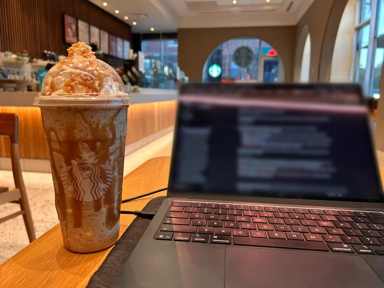 A Venti caramel frappuccino with whipped cream and caramel drizzle next to an open laptop at a Starbucks